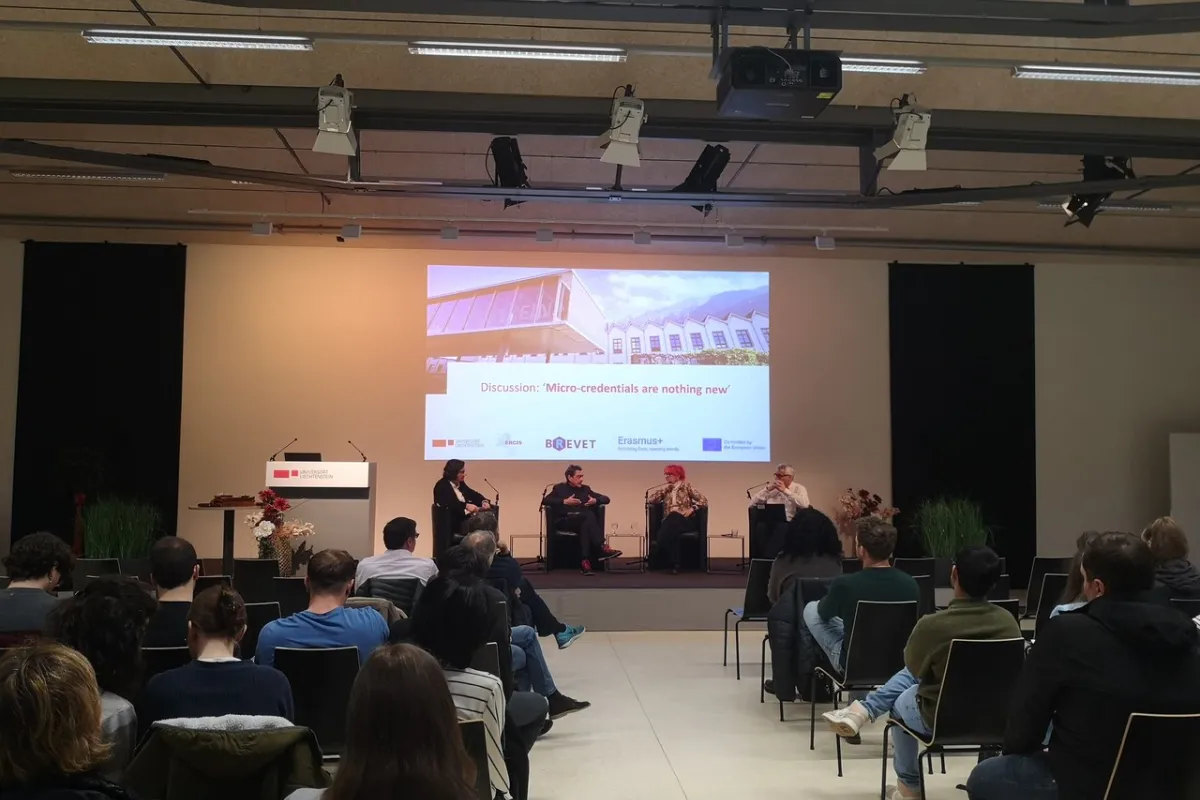 Ein Auditorium mit mehreren Personen, die einer Podiumsdiskussion zuhören. Drei Redner sitzen auf der Bühne vor einer Leinwand mit der Aufschrift „Discussion: 'Micro-credentials are nothing new'“. Die Veranstaltung findet in einem modernen Konferenzraum statt.
