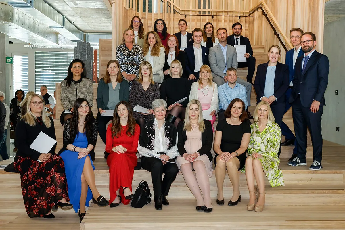 Gruppenfoto von Absolventinnen und Absolventen des Zertifikatsstudiengangs Treuhandwesen der Universität Liechtenstein, die auf einer Treppe sitzen und stehen. Sie halten Zertifikate in den Händen und lächeln in die Kamera. Im Hintergrund ist ein moderner Innenraum zu sehen.