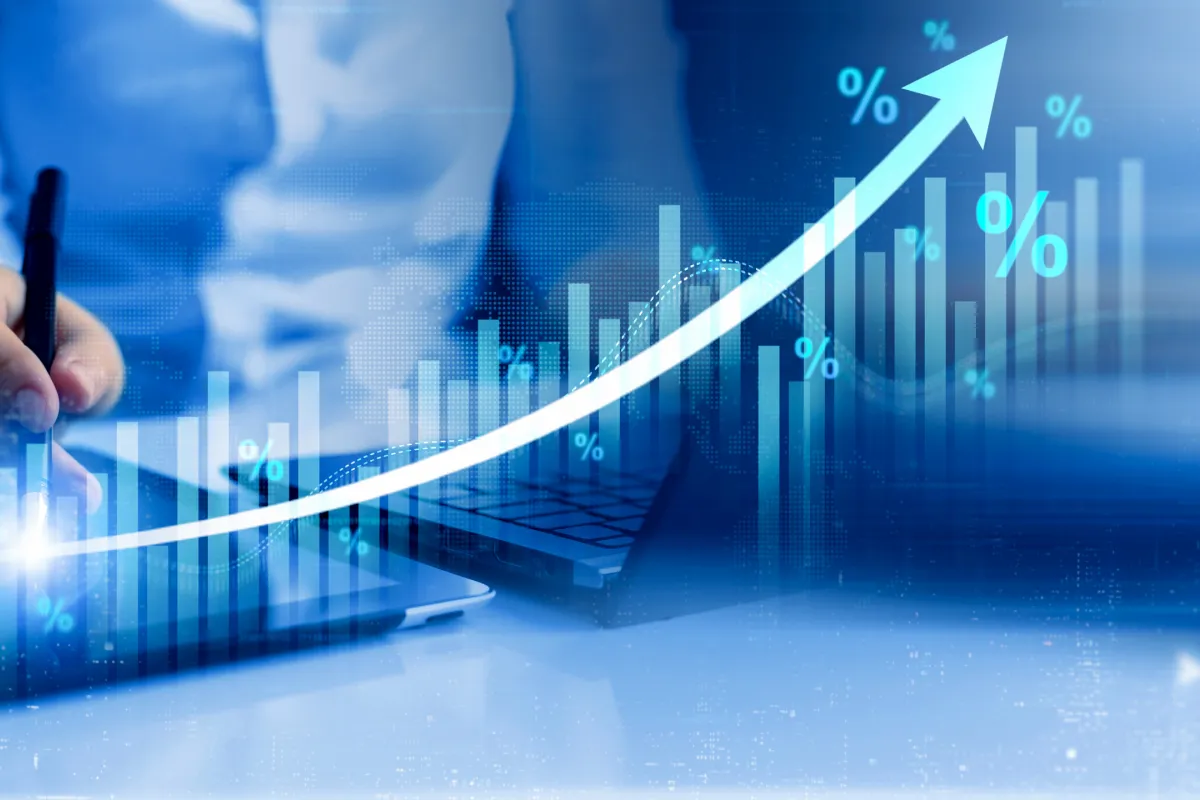 Person analysiert Wirtschaftsdaten an Laptop und Tablet mit steigender Kurve und Prozentzeichen.