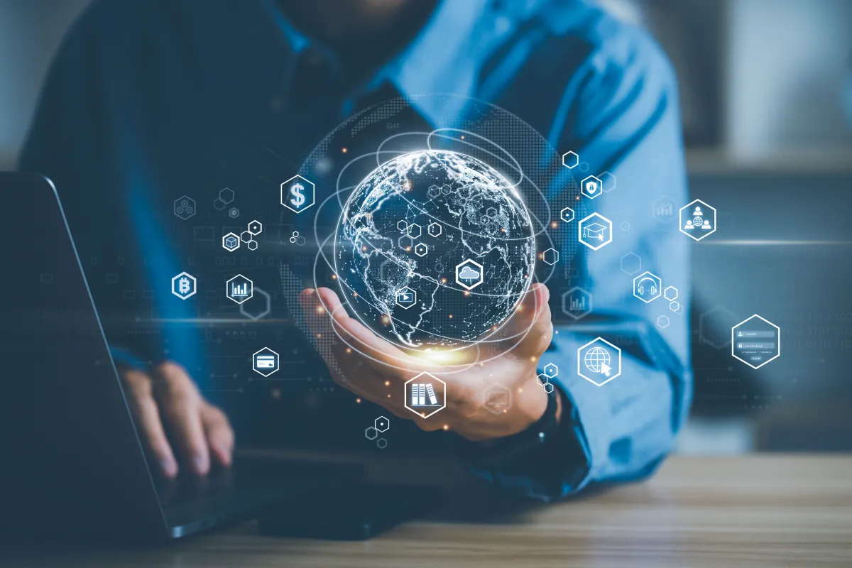 Person hält digitale Weltkugel mit globalen Wirtschaftssymbolen vor Laptop.
