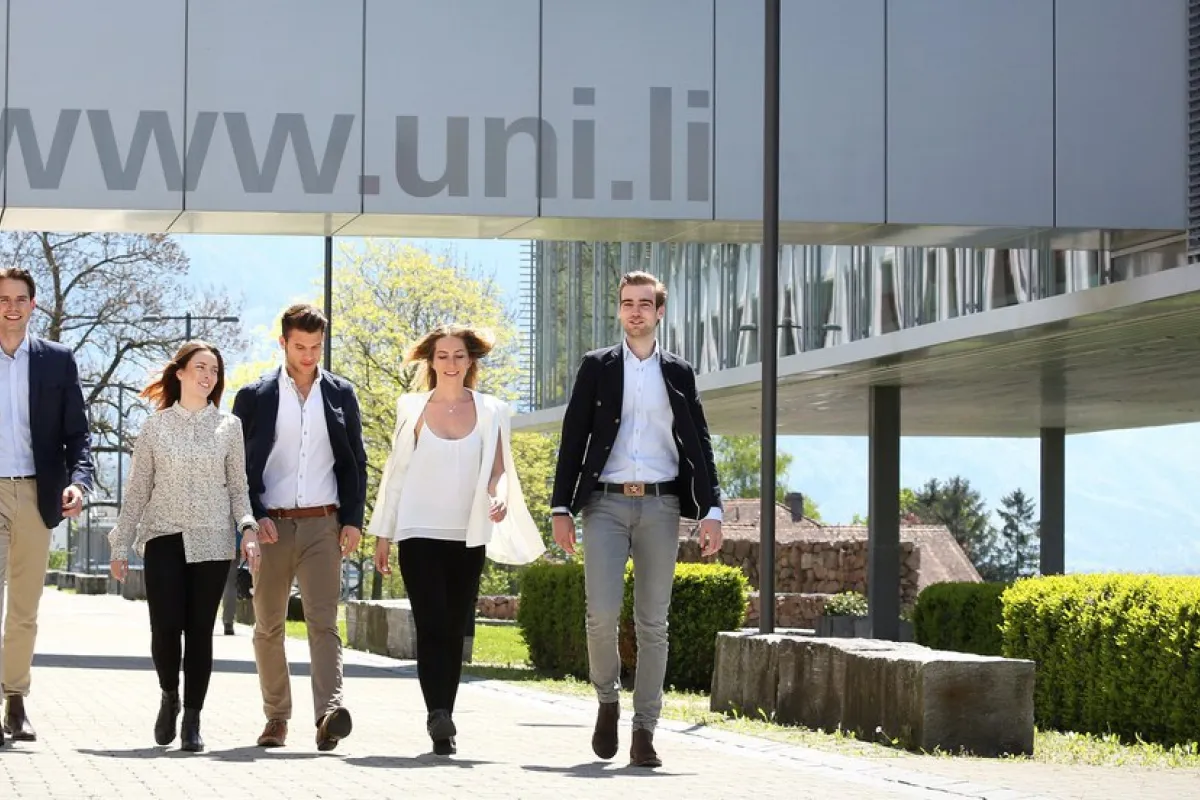 Eine Gruppe von fünf Personen geht lächelnd auf einem sonnigen Campusweg der Universität Liechtenstein entlang, mit der URL "www.uni.li" im Hintergrund.