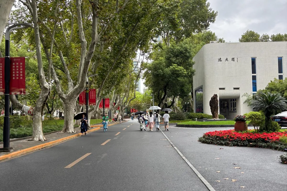 Baumgesäumte Allee auf einem Campus mit Spaziergängern, roten Bannern und einem modernen Gebäude am Straßenrand.