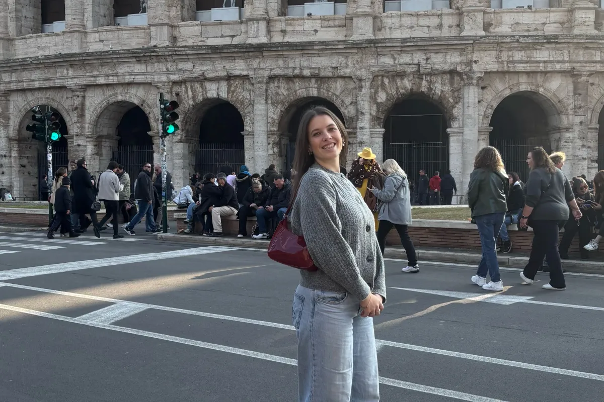 Frau steht lächelnd vor dem Kolosseum, während im Hintergrund Touristen an der Straße entlanggehen.
