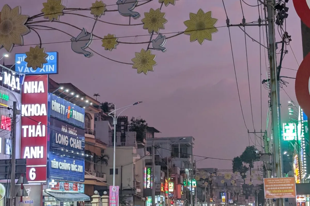 Belebte Straße bei Dämmerung mit vielen Motorrollern, leuchtenden Werbeschildern und dekorativen Lichtern über der Fahrbahn.