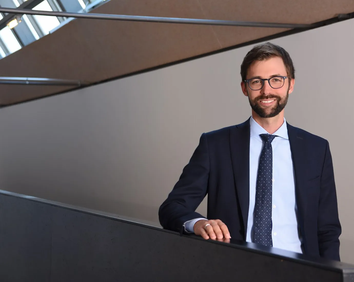 Ein Mann im Anzug steht lächelnd in einem modernen Gebäude mit schrägen Fenstern.