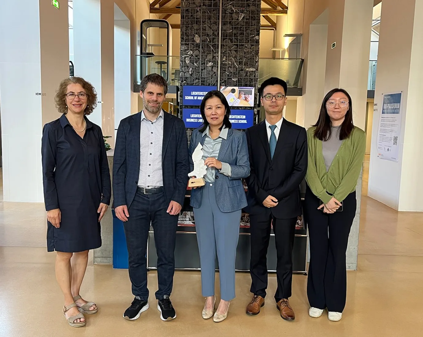 Eine Gruppe von fünf Personen steht in einem modernen Gebäude der Universität Liechtenstein. Sie lächeln in die Kamera. Im Hintergrund sind Informationsschilder der Universität zu sehen.