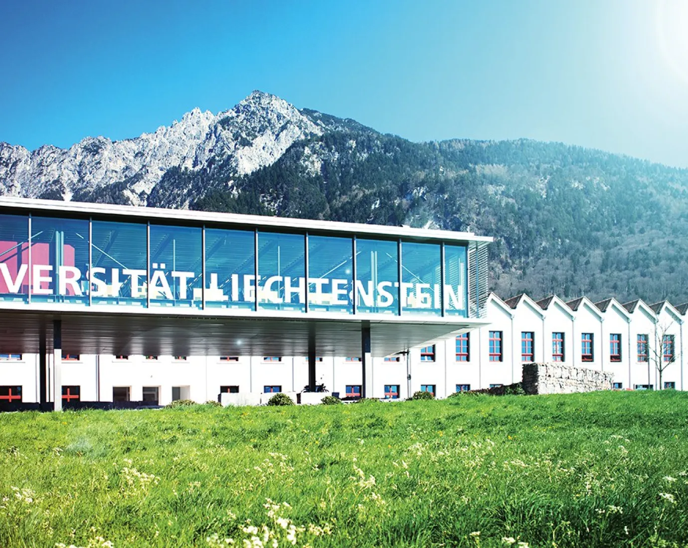 Außenansicht der Universität Liechtenstein mit modernem Glasgebäude und Bergkulisse im Hintergrund, umgeben von grüner Wiese.