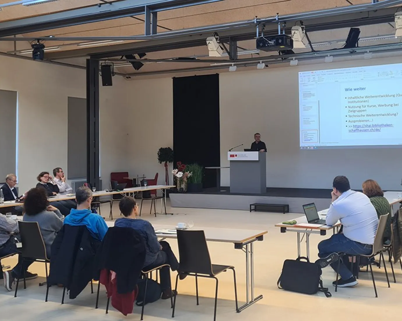 Eine Gruppe von etwa 20 Personen sitzt in einem Auditorium der Universität Liechtenstein, während ein Vortragender an einem Rednerpult spricht. Auf einer großen Leinwand ist eine Präsentation mit dem Titel "Wie weiter" zu sehen.