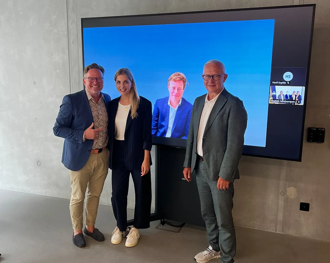 Drei Personen stehen vor einem Bildschirm, der zwei weitere Personen in einem virtuellen Meeting zeigt. Die Szene befindet sich in einem modernen Raum mit Betonwänden.