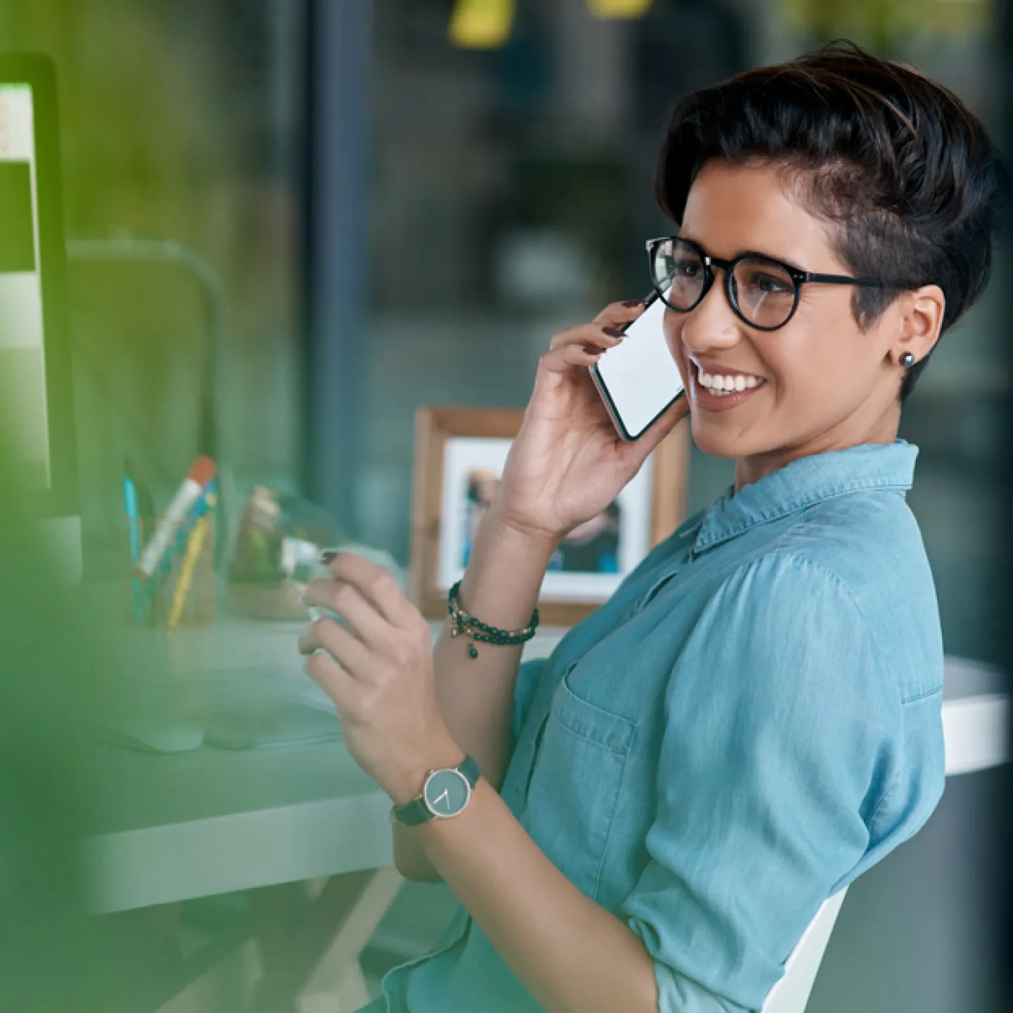 Eine junge Frau mit Kurzhaarfrisur und Brille sitzt an einem Schreibtisch und telefoniert lächelnd mit einem Smartphone. Sie trägt ein blaues Hemd und schaut entspannt aus dem Fenster. Vor ihr steht ein Computerbildschirm, und im Hintergrund sind Büroaccessoires und persönliche Gegenstände zu sehen. Die Atmosphäre ist modern und professionell.