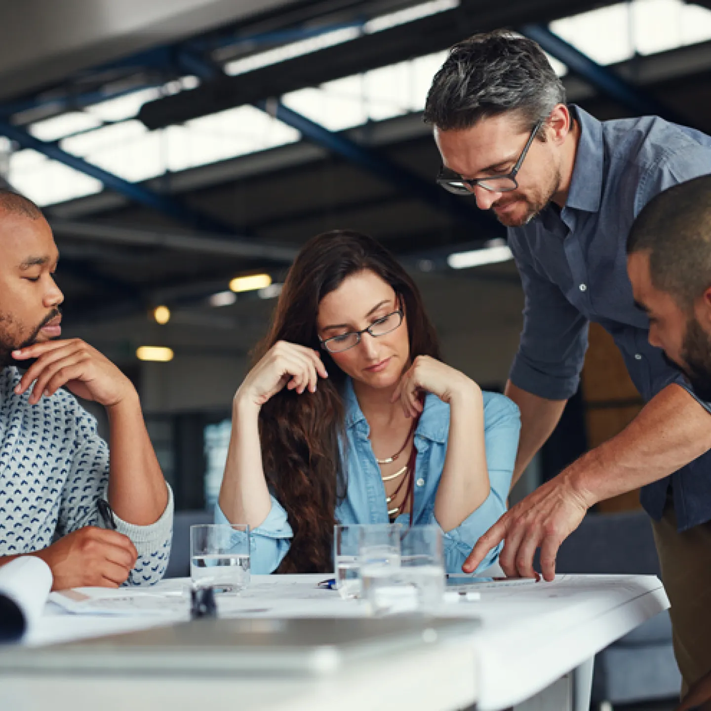Eine Gruppe von vier Menschen, die um einen Tisch in einem modernen Büro sitzen und gemeinsam an einem Projekt arbeiten. Zwei Männer, eine Frau und ein weiterer Mann, der auf das Dokument zeigt, sind in eine Diskussion vertieft. Vor ihnen liegen Pläne und Dokumente, auf dem Tisch stehen Gläser mit Wasser. Die Atmosphäre wirkt konzentriert und kollaborativ.