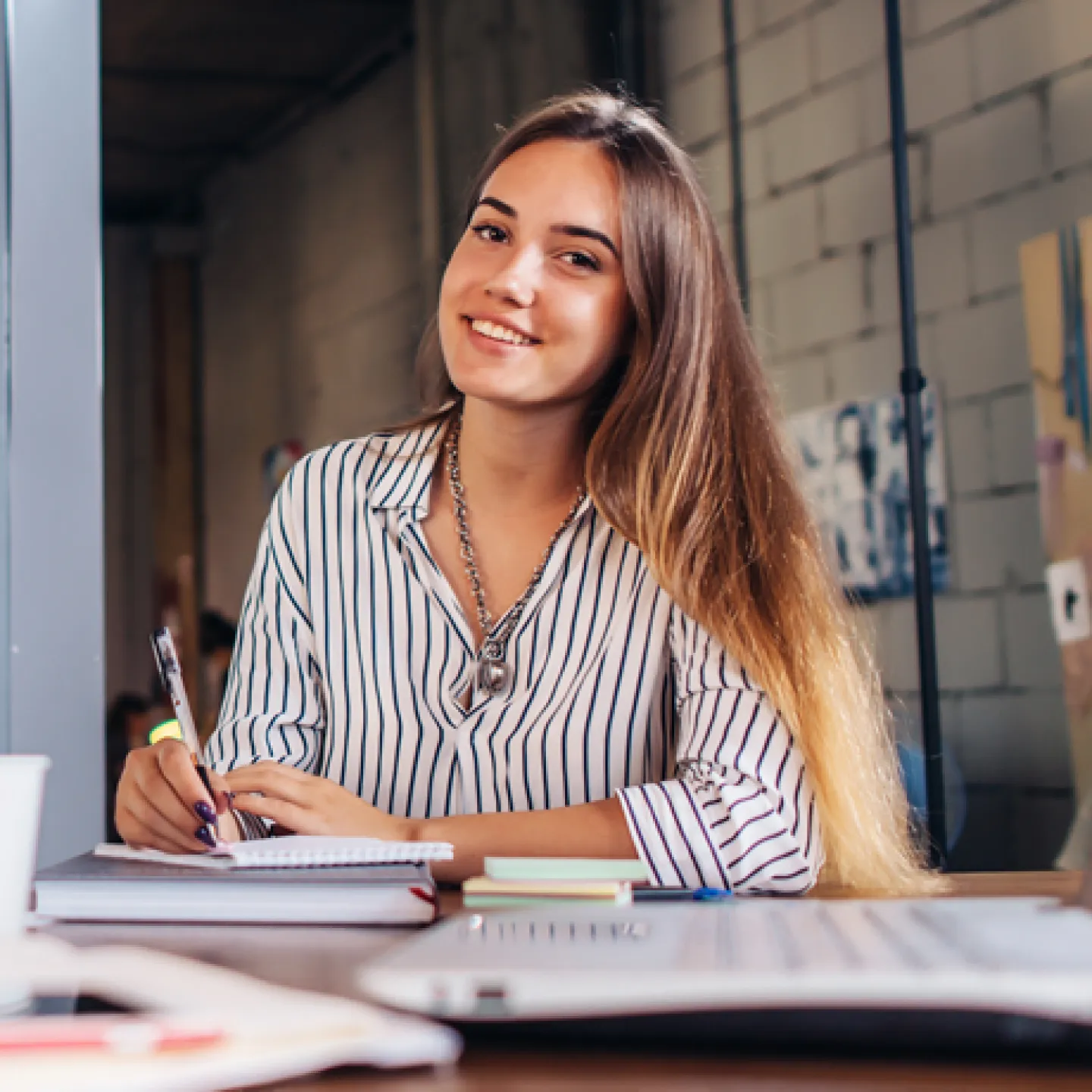 Eine junge Frau sitzt an einem Schreibtisch mit einem Notizbuch und lächelt freundlich in die Kamera. Sie trägt ein gestreiftes Hemd, und im Hintergrund ist eine kreative Büroumgebung zu sehen. Der Schreibtisch ist mit Laptop, Notizen und einer Kaffeetasse bedeckt, während sie einen Stift in der Hand hält, bereit zu schreiben.