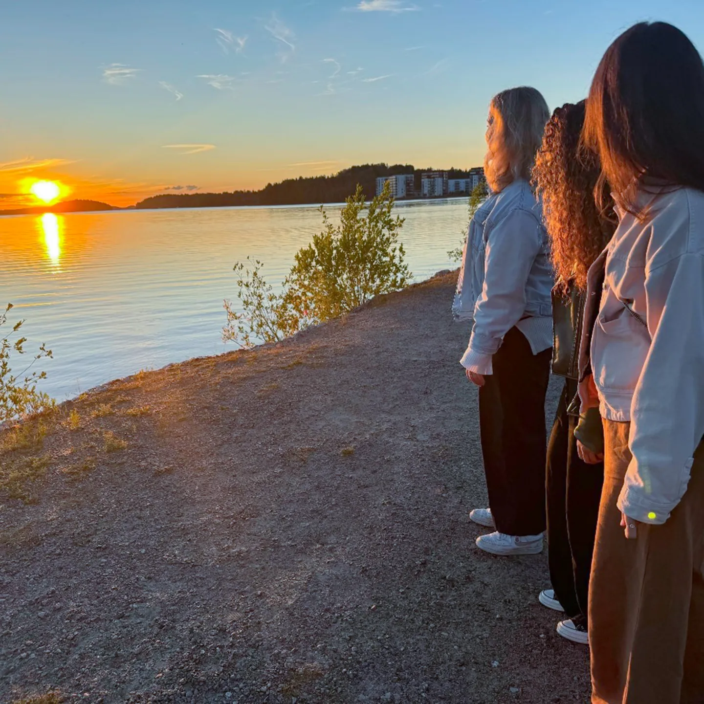 Drei Studentinnen der Universität Liechtenstein beobachten gemeinsam den Sonnenuntergang an einem ruhigen Seeufer.