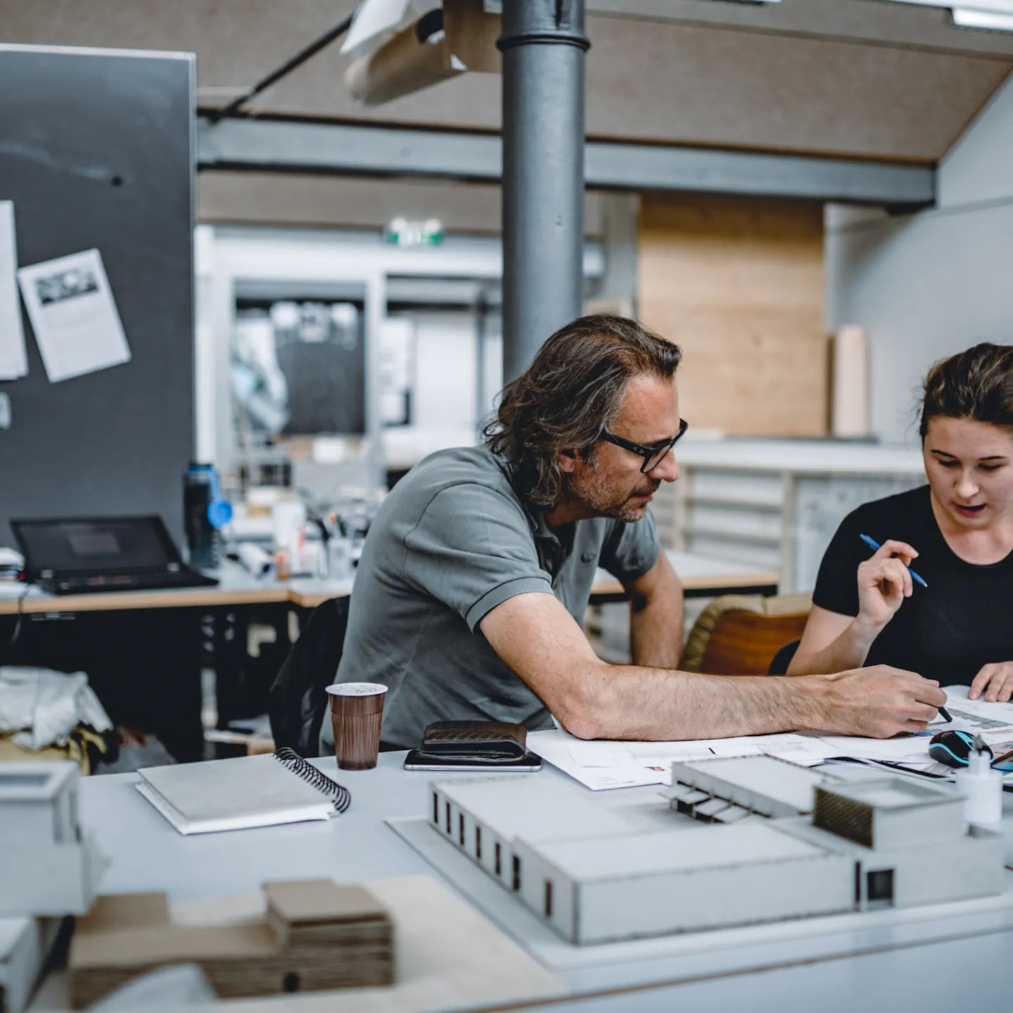 Dozent unterstützt eine Studentin bei der Projektarbeit im Architekturatelier der Universität Liechtenstein, umgeben von Modellen und Unterlagen.