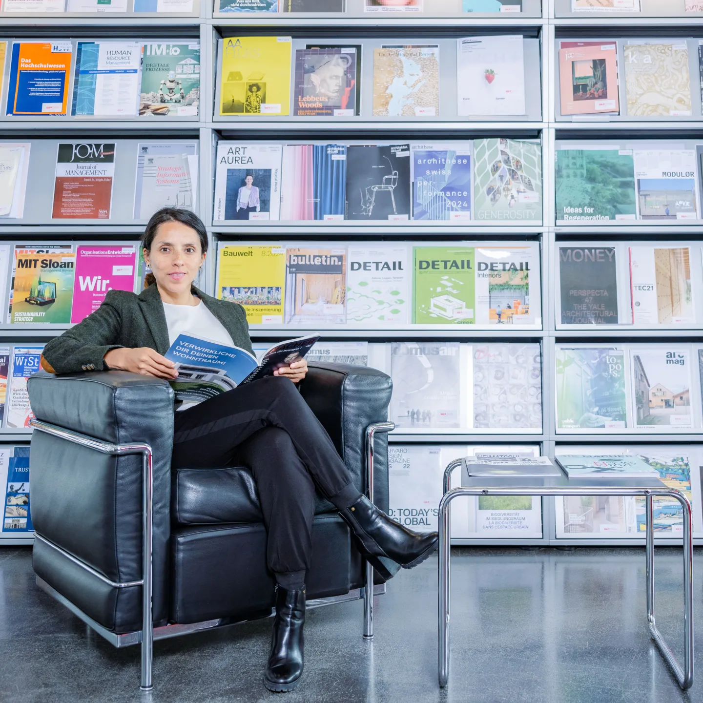 Frau sitzt entspannt auf einem schwarzen Ledersessel in der Bibliothek der Universität Liechtenstein und liest in einer Fachzeitschrift, im Hintergrund ein Regal mit bunten Magazincovern.