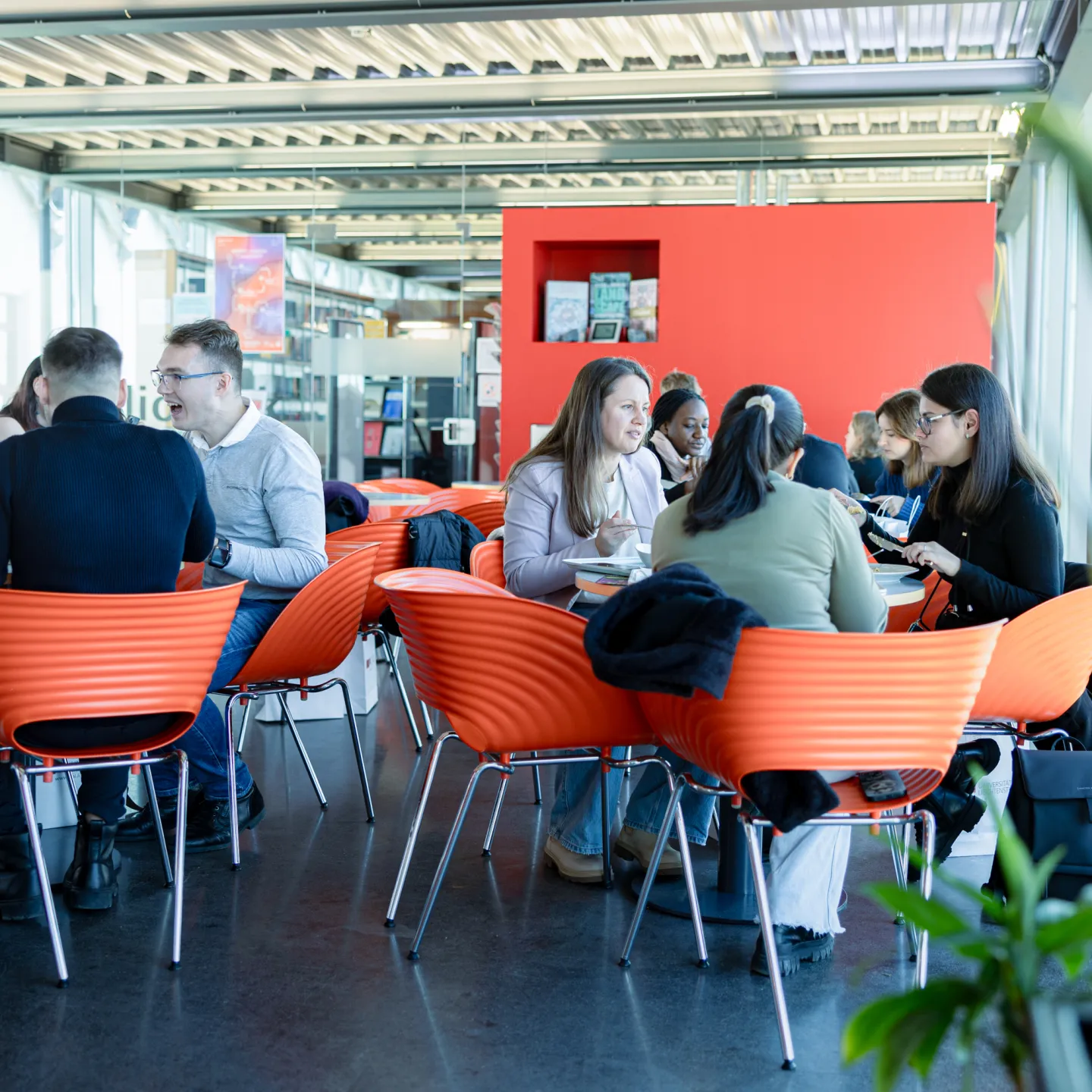 Lebendige Szene in der Universitätscafeteria mit mehreren Gruppen Studierender beim gemeinsamen Mittagessen und Gesprächen.