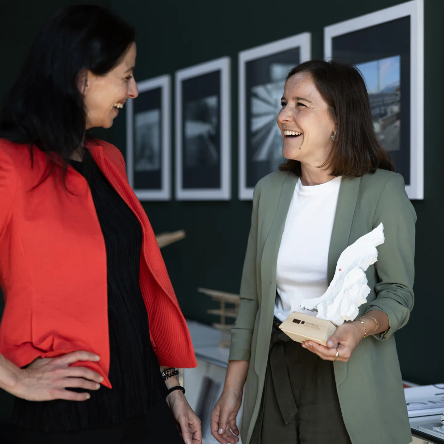 Zwei Frauen im Gespräch vor einer Bilderwand an der Universität Liechtenstein, eine hält ein Architekturmodell.