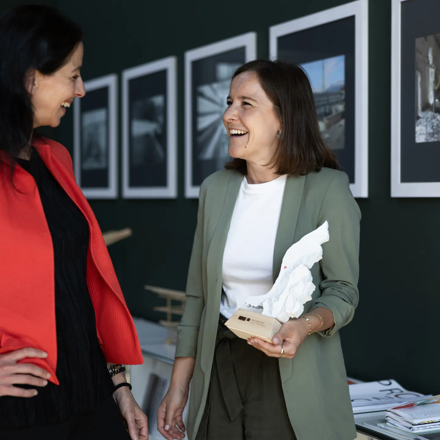 Zwei Frauen im Gespräch vor einer Bilderwand an der Universität Liechtenstein, eine hält ein Architekturmodell.