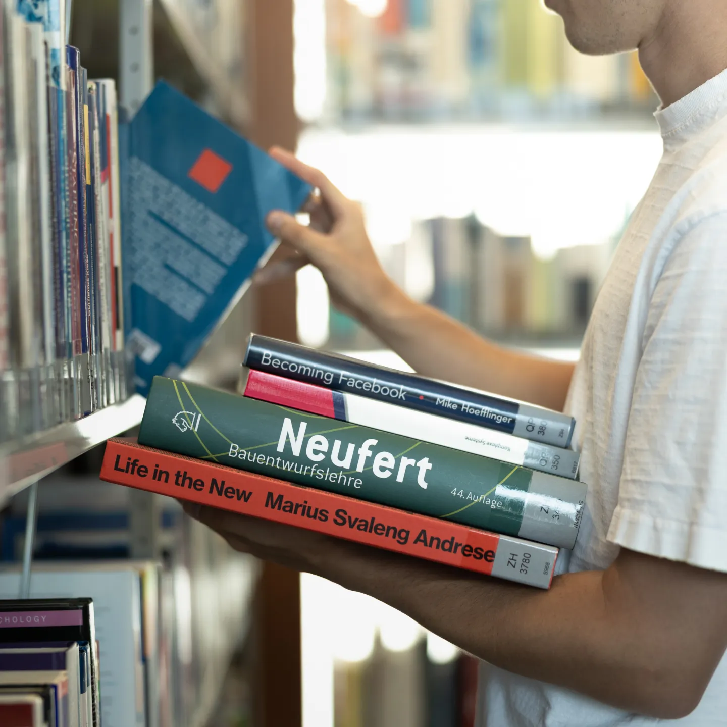 Student wählt Fachliteratur aus dem Bücherregal der Universitätsbibliothek Liechtenstein, mehrere Bücher im Arm.