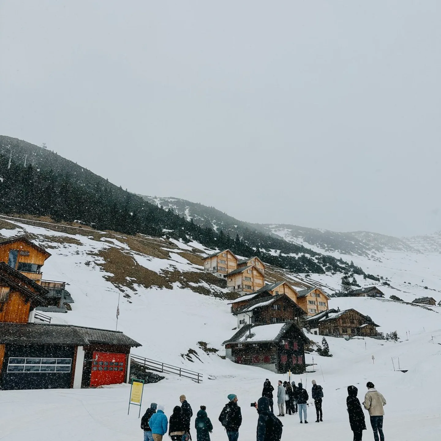 Eine Gruppe von Menschen steht in einer verschneiten Berglandschaft vor traditionellen Holzhäusern. Im Hintergrund sind schneebedeckte Hügel und Nadelbäume zu sehen. Die Szene vermittelt eine winterliche Atmosphäre und zeigt eine Verbindung von Architektur und Natur.