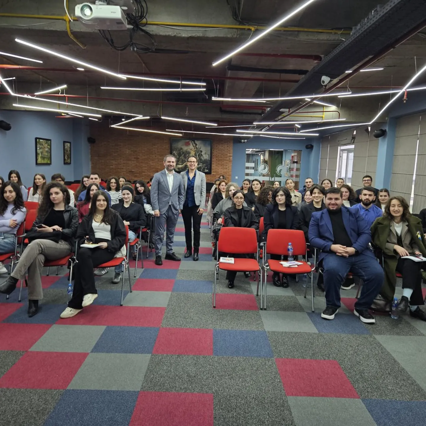 Seminarraum mit Studierenden und Dozenten, die an einem Cybercrime-Seminar teilnehmen. Die Teilnehmenden sitzen auf roten Stühlen in einem modernen Raum mit geometrischem Teppichmuster und futuristischer Beleuchtung.