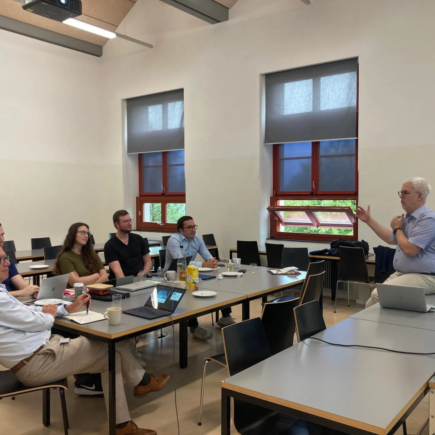 Ein Dozent hält einen Vortrag vor einer kleinen Gruppe von Personen in einem Seminarraum. Die Teilnehmenden sitzen an Tischen mit Laptops und Notizbüchern. Im Hintergrund sind Fenster und eine Leinwand zu sehen.