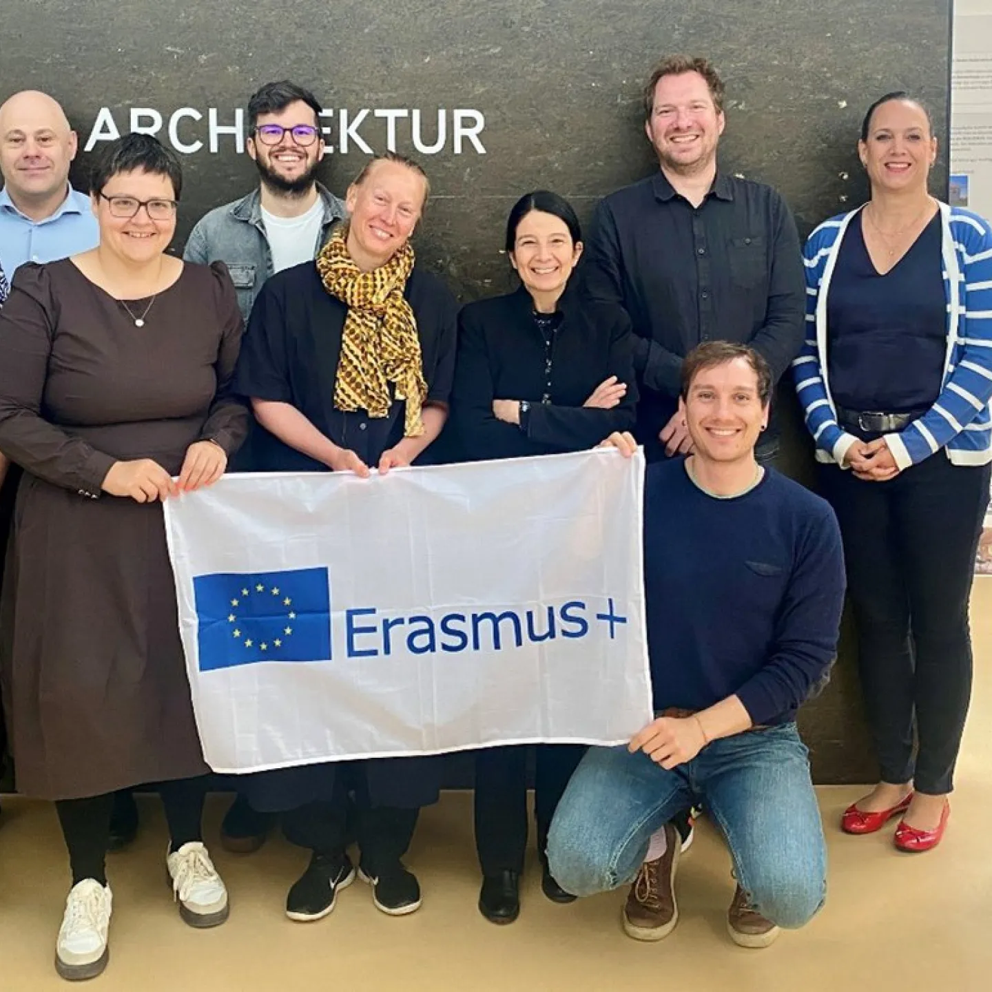 Gruppe von zehn Personen steht vor einer Wand mit dem Schriftzug "Architektur" und hält ein Erasmus+ Banner. Sie lächeln in die Kamera.
