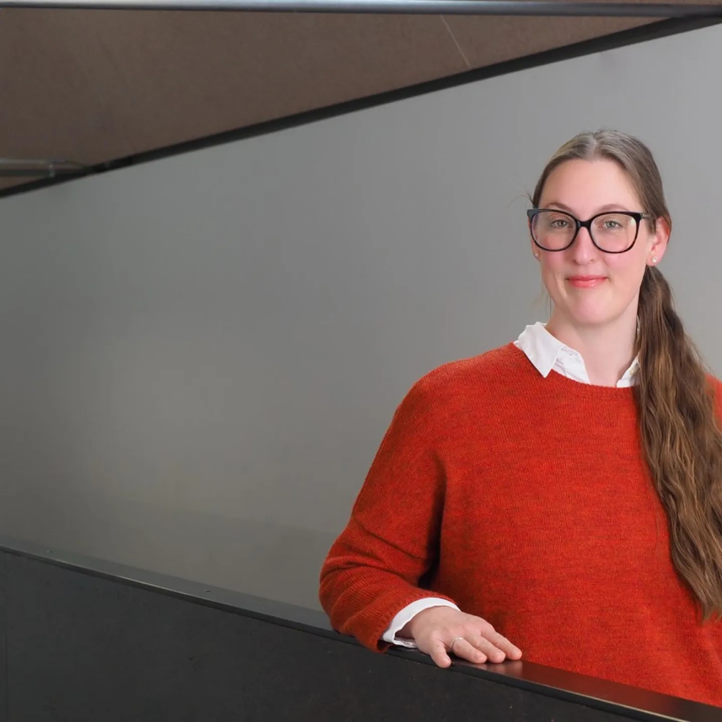 Eine Person mit Brille und langem Haar steht in einem modernen Gebäude und trägt einen roten Pullover. Sie lächelt leicht und lehnt sich an ein Geländer.