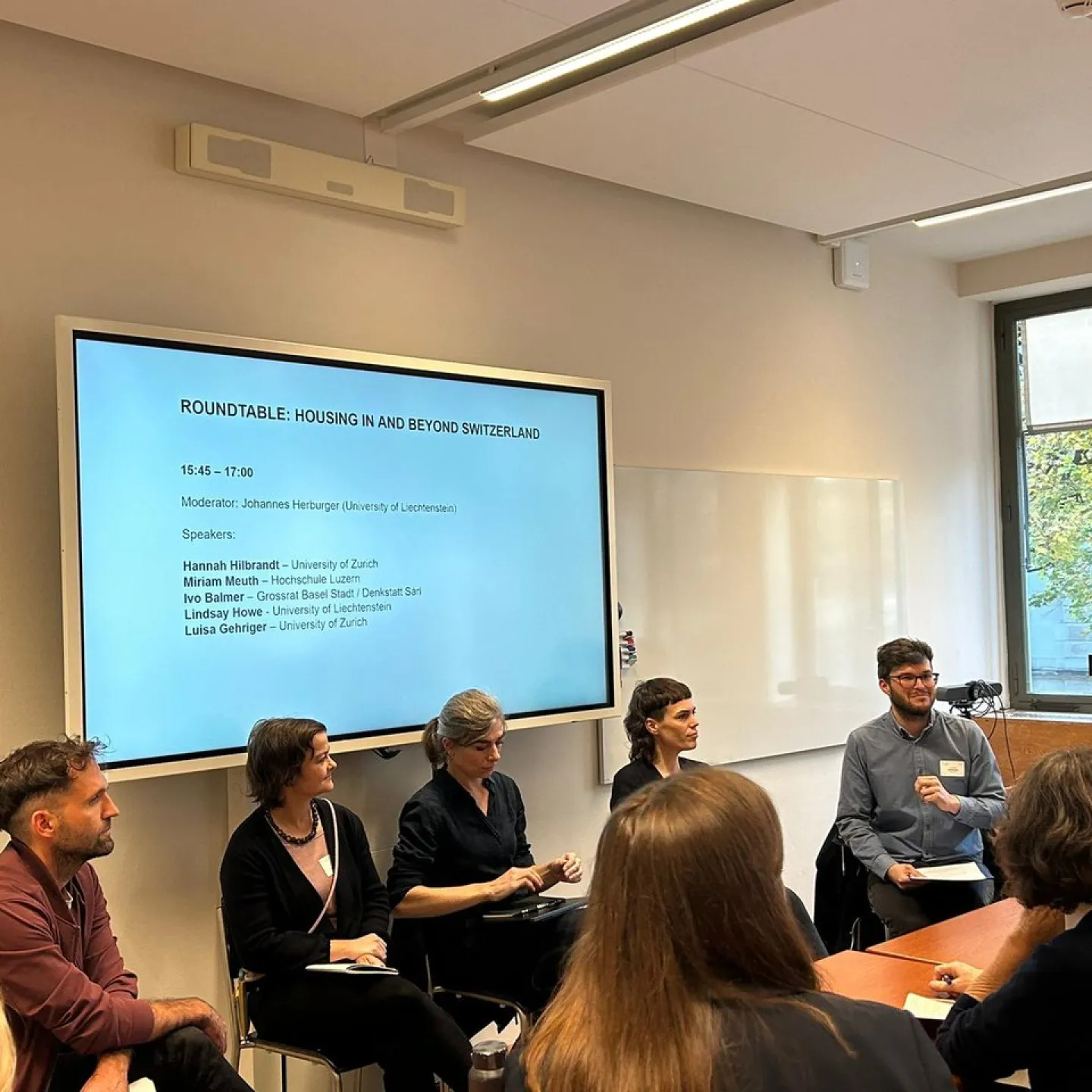 Eine Gruppe von Personen sitzt in einem Seminarraum und diskutiert bei einer Podiumsdiskussion zum Thema "Housing in and Beyond Switzerland". Im Hintergrund ist ein Bildschirm mit den Namen der Teilnehmenden und dem Moderator zu sehen.