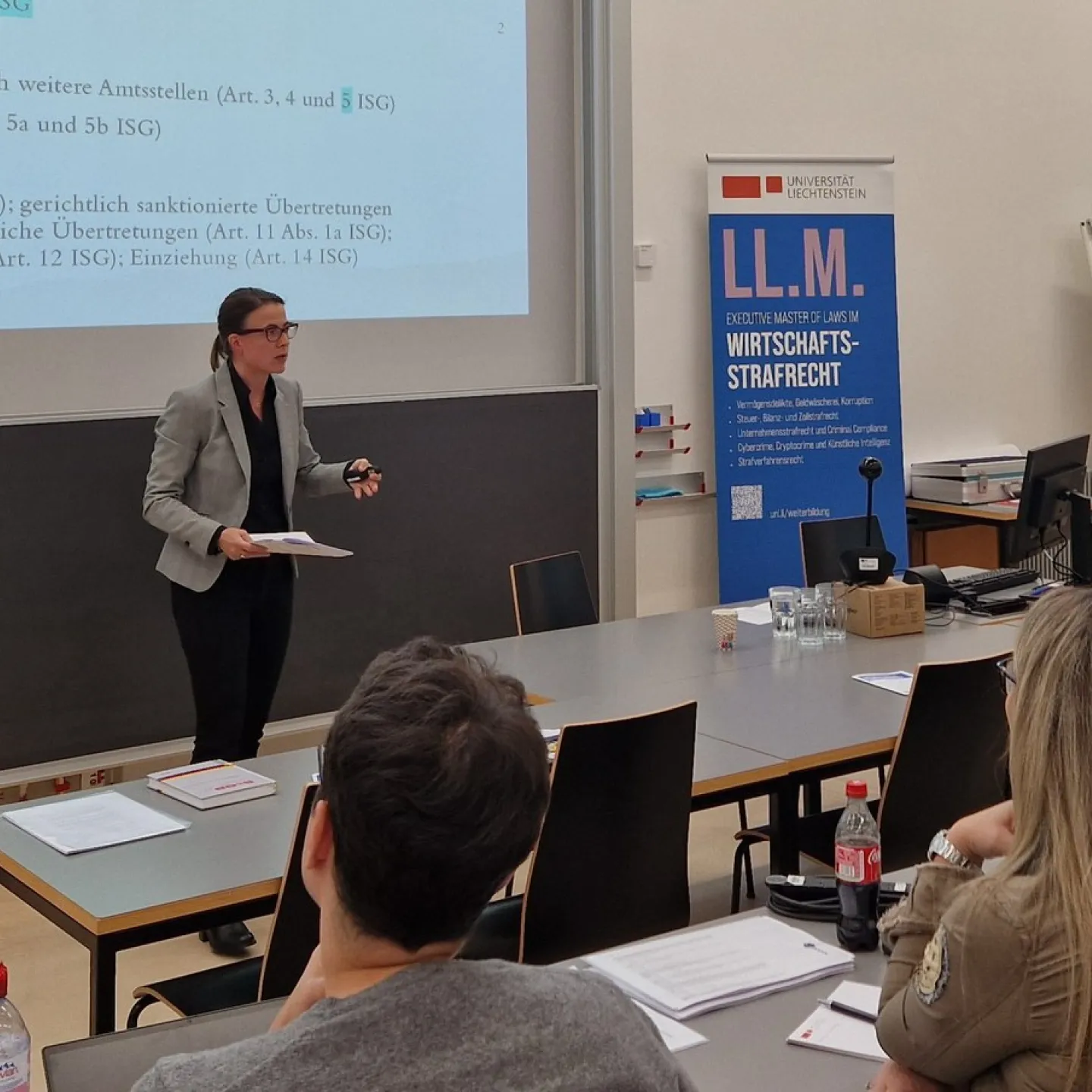Eine Person hält einen Vortrag in einem Seminarraum der Universität Liechtenstein. Im Hintergrund ist eine Präsentationsfolie zu sehen. Rechts steht ein Roll-Up mit der Aufschrift "LL.M. Wirtschafts-Strafrecht". Teilnehmer sitzen an Tischen und hören zu.