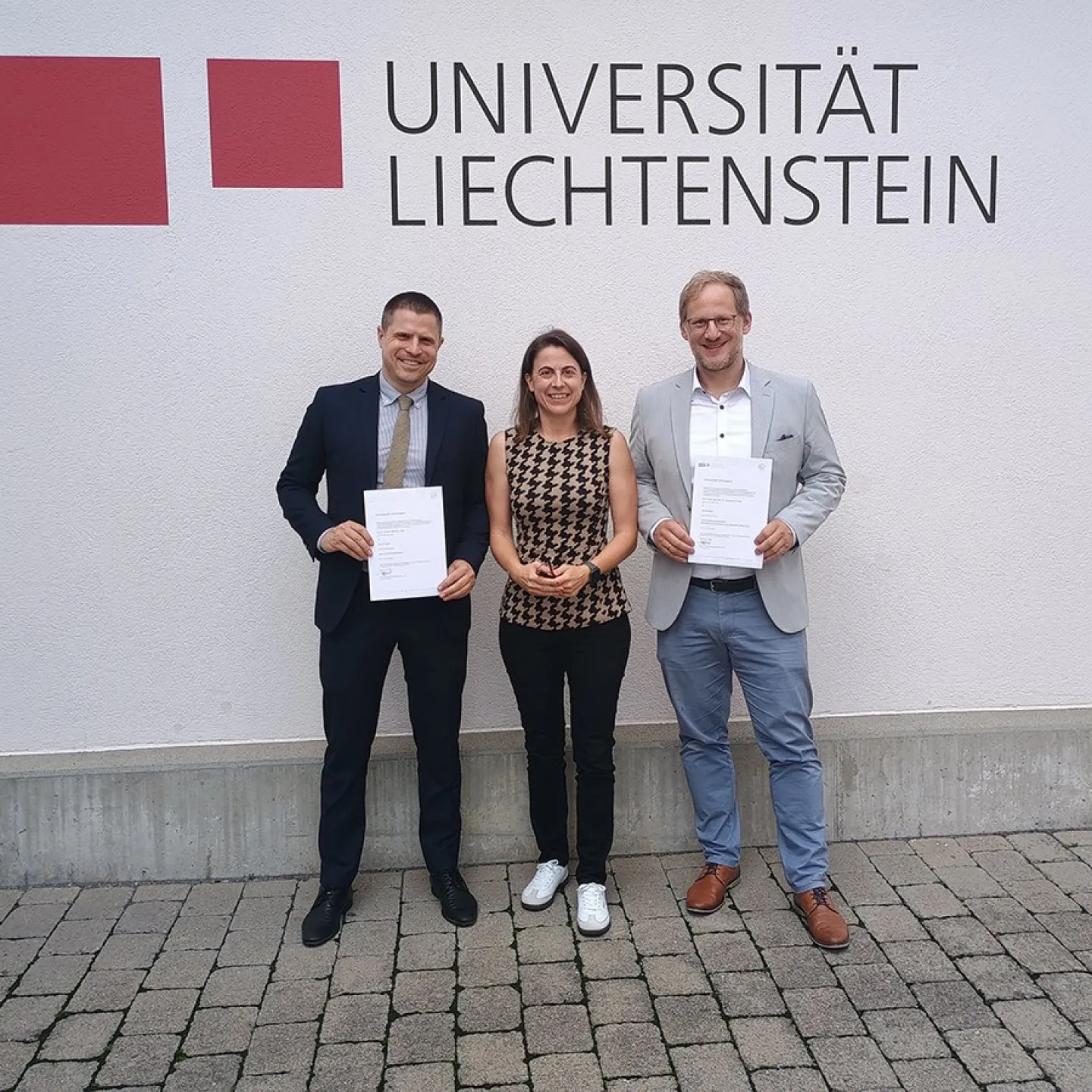 Drei Personen stehen vor einer Wand mit dem Logo der Universität Liechtenstein. Zwei von ihnen halten Zertifikate in den Händen.