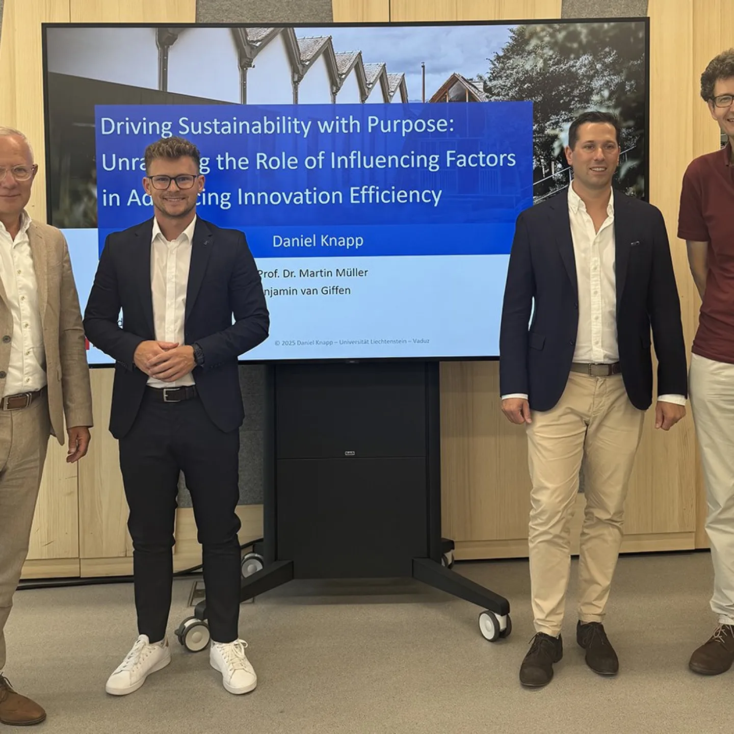 Vier Männer stehen vor einem Bildschirm, der eine Präsentation mit dem Titel „Driving Sustainability with Purpose“ zeigt. Sie tragen Business-Kleidung und lächeln in die Kamera. Der Hintergrund ist ein moderner Innenraum.