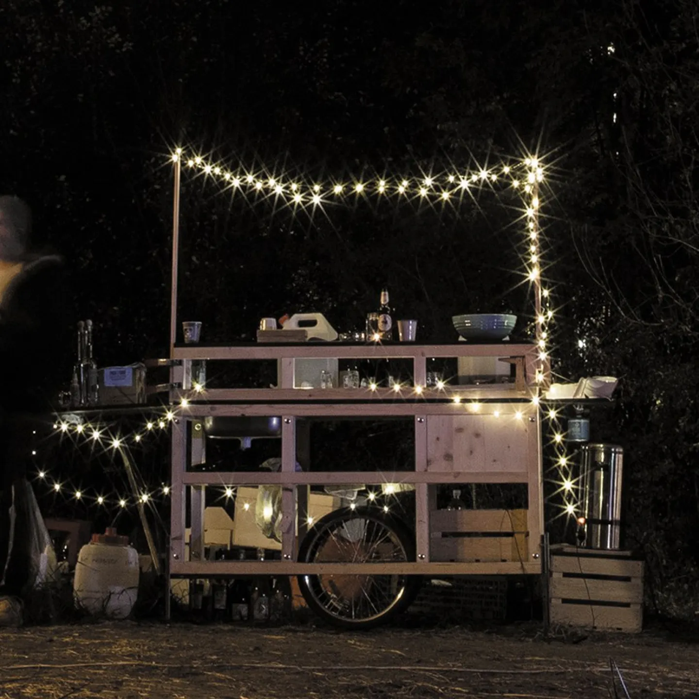 Eine mobile Holzbar, dekoriert mit Lichterketten, steht im Freien bei Nacht. Mehrere Personen stehen links neben der Bar und unterhalten sich. Die Szene vermittelt eine gemütliche und einladende Atmosphäre.