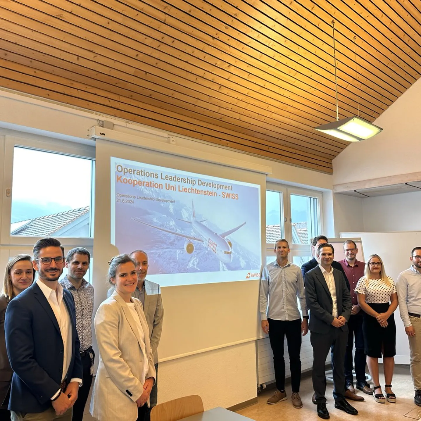 Eine Gruppe von Menschen steht lächelnd in einem Seminarraum vor einer Leinwand, die eine Präsentation zum Operations Leadership Development-Programm der Universität Liechtenstein und SWISS zeigt.