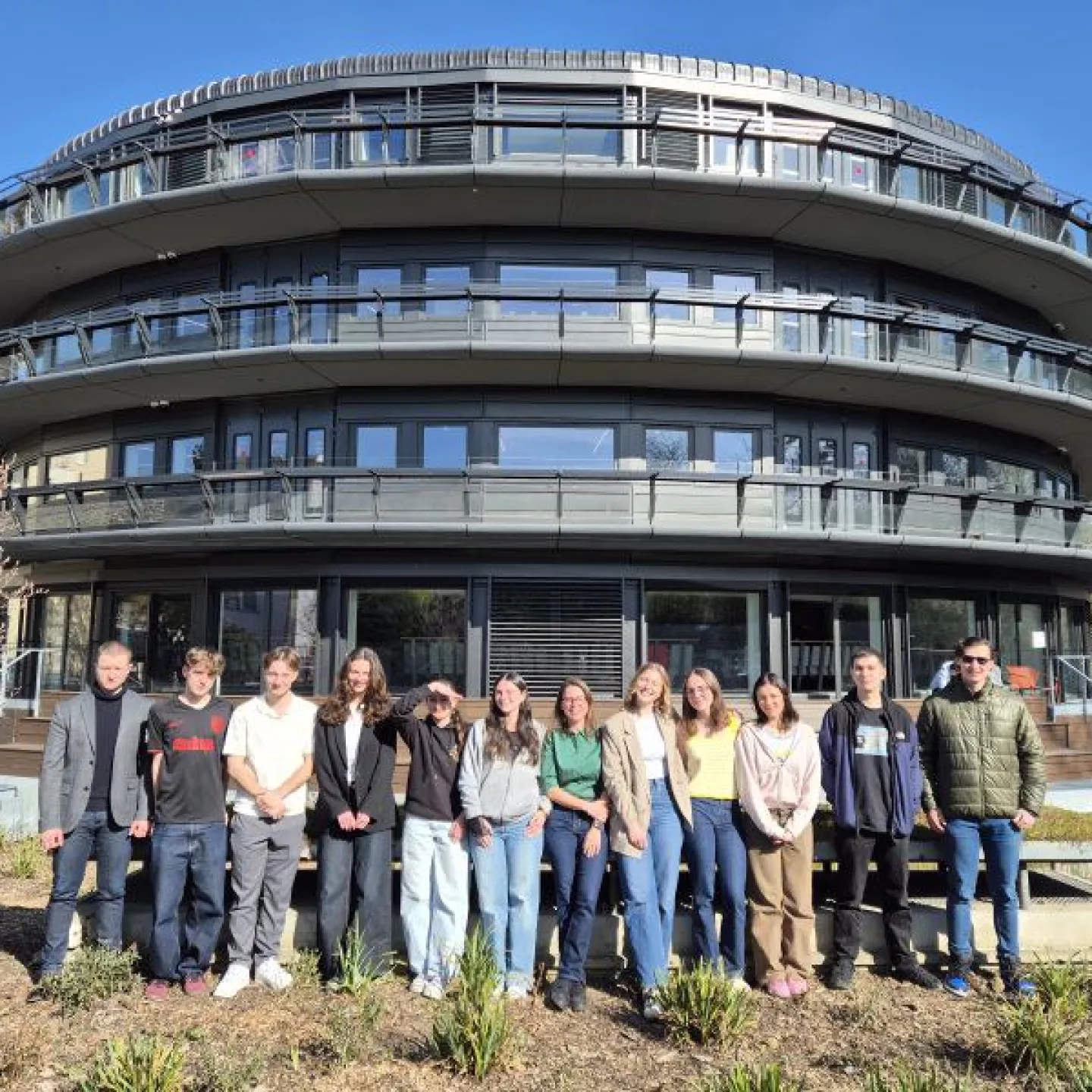 Gruppe von Studierenden und einer Lehrperson vor einem modernen, runden Universitätsgebäude bei sonnigem Wetter.