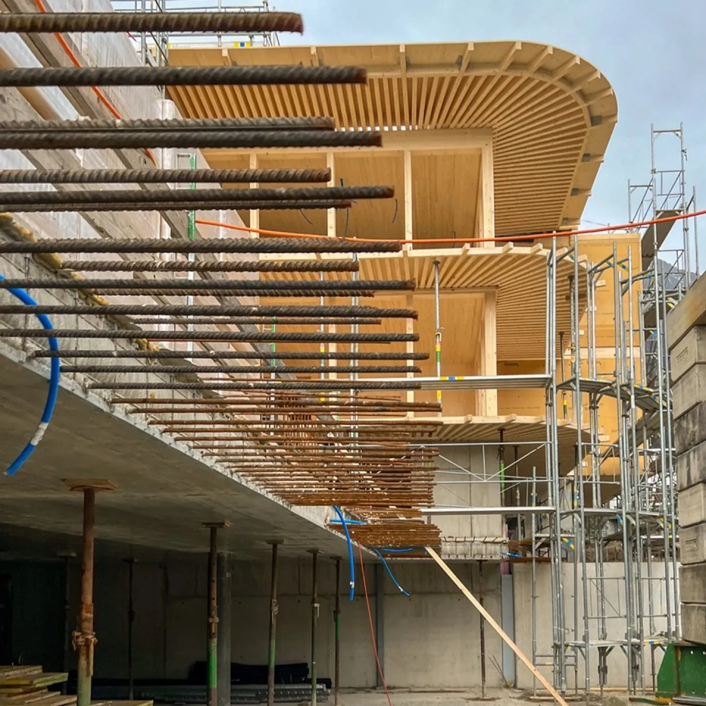 Baustelle eines modernen Gebäudes mit sichtbaren Stahlträgern und Holzverkleidung, umgeben von Gerüsten, unter bewölktem Himmel.