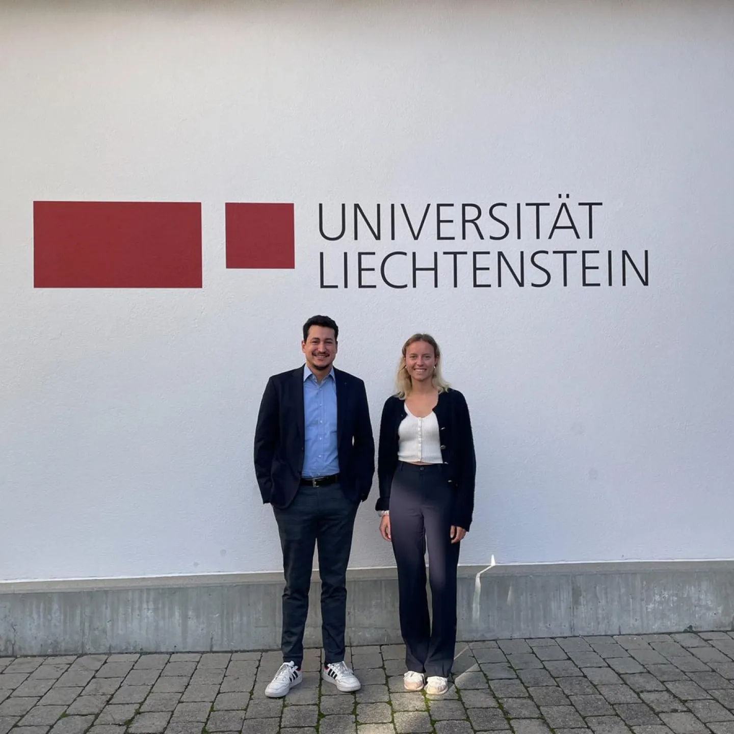 Zwei Personen stehen vor einer Wand mit dem Logo der Universität Liechtenstein. Sie lächeln in die Kamera.