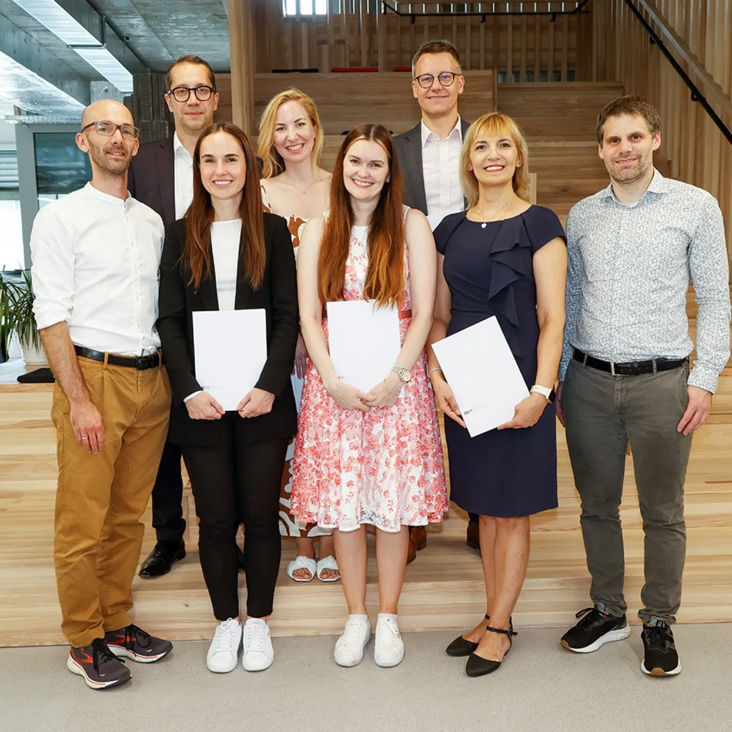 Eine Gruppe von acht Personen steht lächelnd in einem modernen Innenraum. Drei Personen in der vorderen Reihe halten Zertifikate in den Händen. Die Umgebung wirkt hell und einladend mit Holzelementen und Pflanzen.
