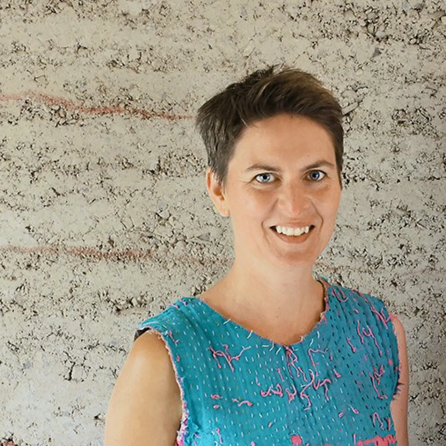 Eine lächelnde Person steht vor einer strukturierten Lehmwand. Sie trägt ein blaues Oberteil mit rosa Akzenten.