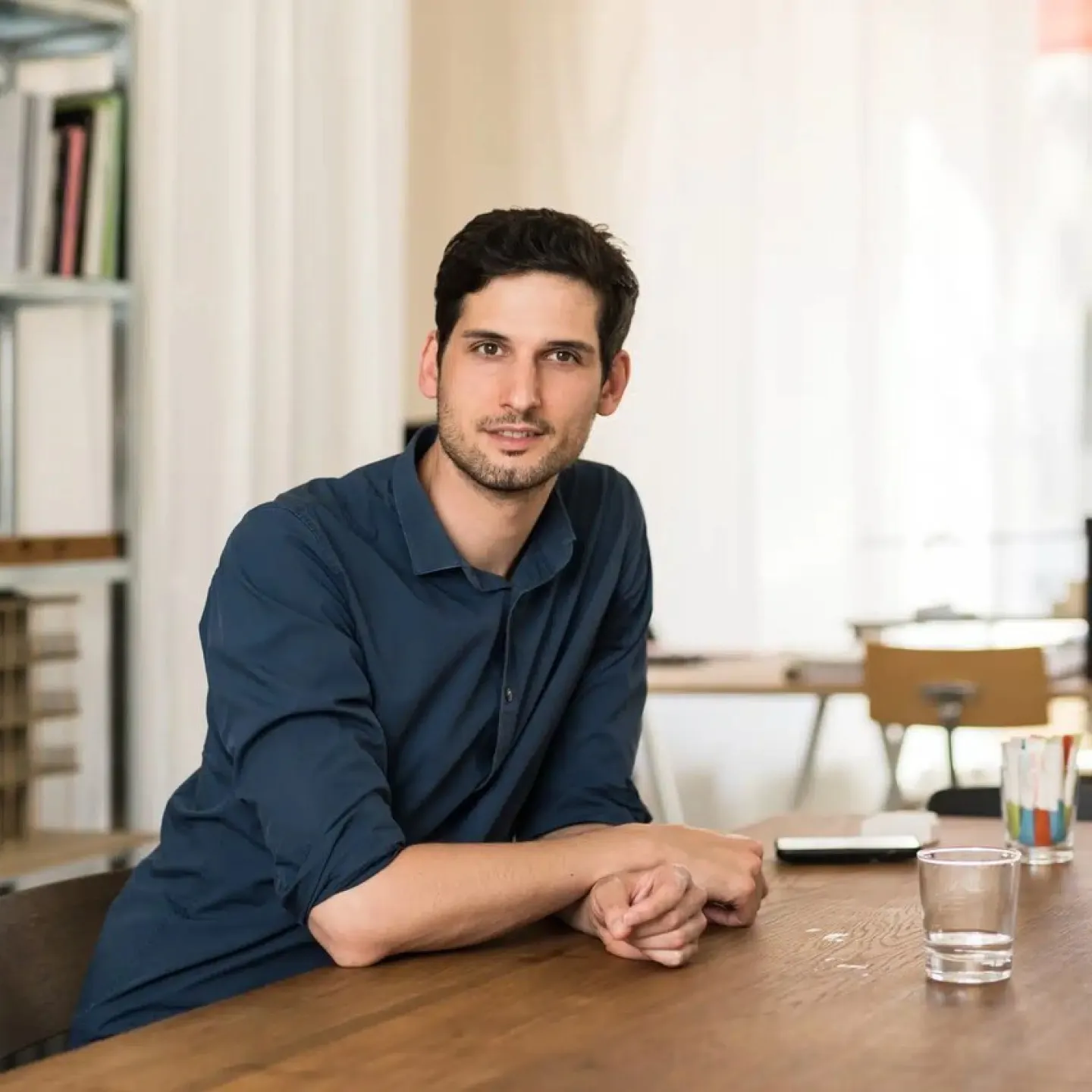 Ein Mann mit blauem Hemd sitzt an einem Tisch und hat ein Glas Wasser vor sich stehen.