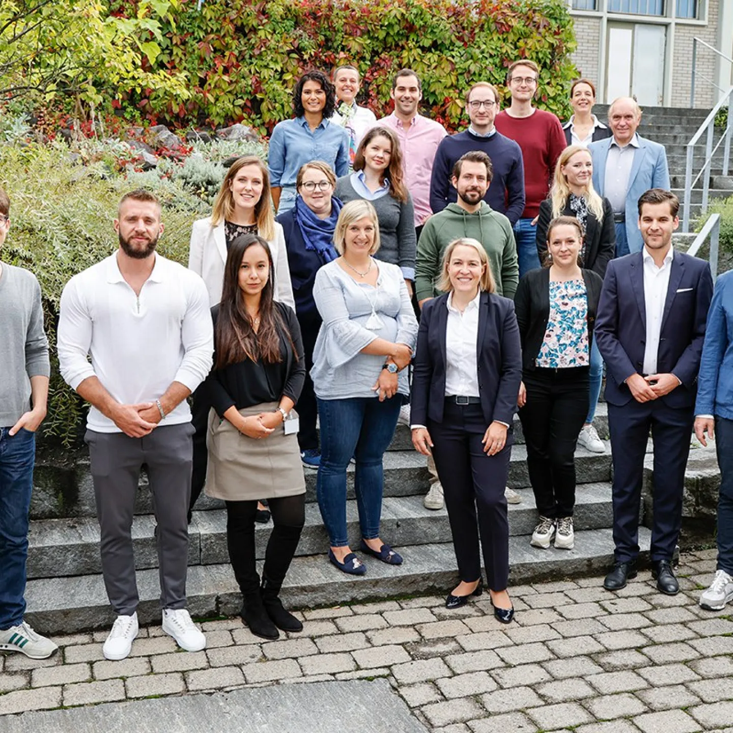 Eine Gruppe von Studierenden und Dozenten des Executive Master of Laws (LL.M.) im Gesellschafts-, Stiftungs- und Trustrecht steht auf einer Treppe im Freien. Sie lächeln in die Kamera, umgeben von grüner Vegetation und einem Gebäude im Hintergrund.