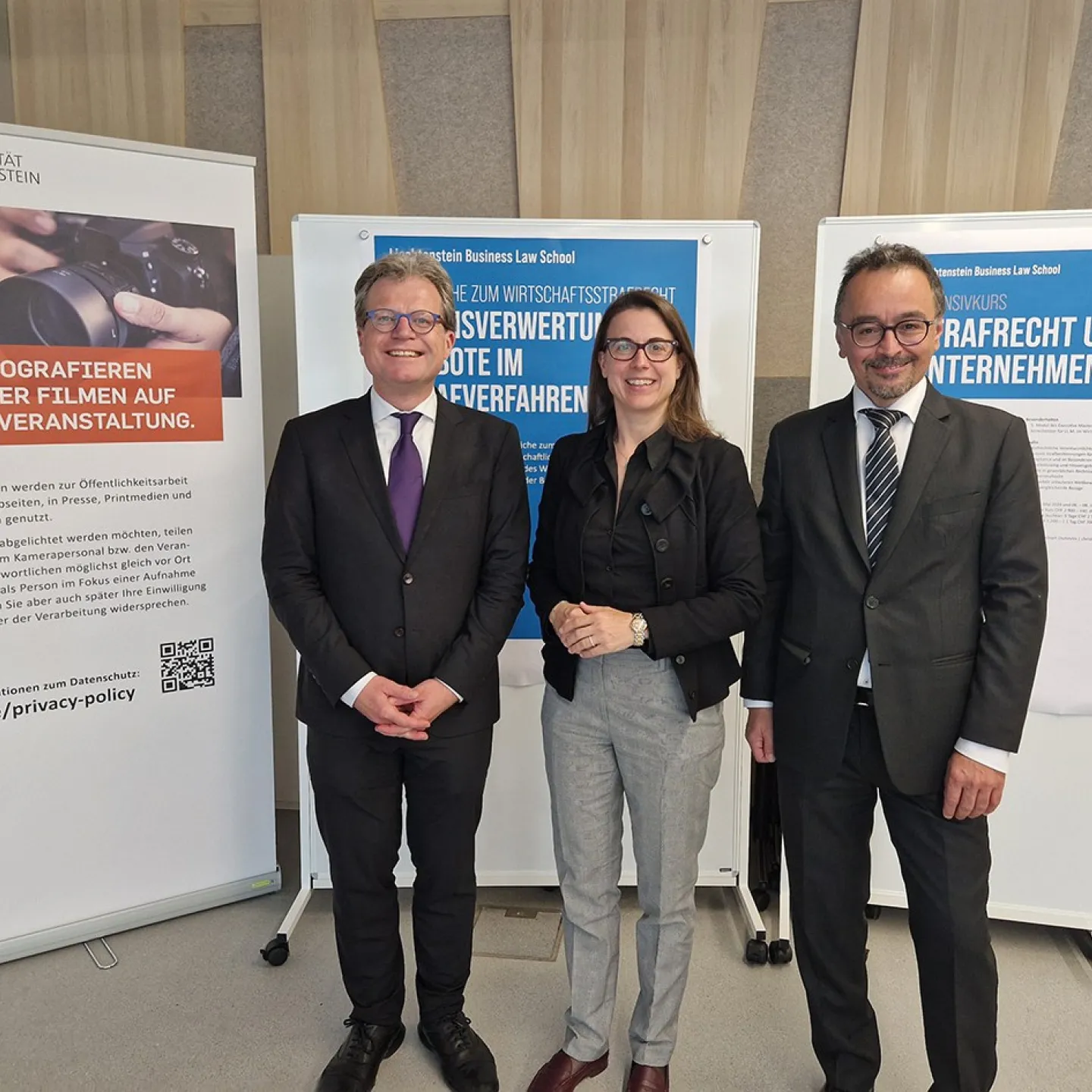Drei Personen stehen lächelnd vor Informationsplakaten der Universität Liechtenstein. Die Plakate thematisieren Wirtschaftsstrafrecht und Beweisverwertungsverbote. Die Personen tragen formelle Kleidung.