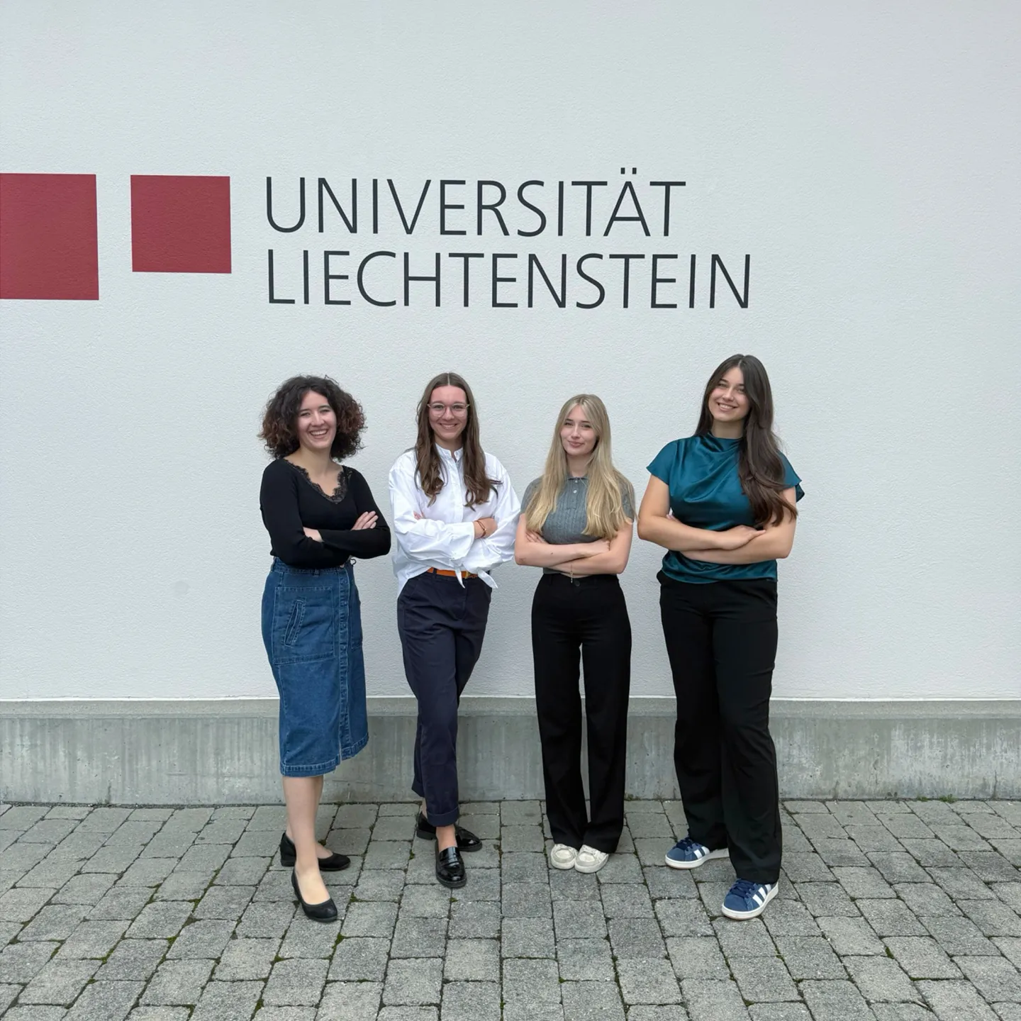 Vier Studentinnen stehen vor der Fassade der Universität Liechtenstein mit Logo und Schriftzug, lächelnd und mit verschränkten Armen, Symbol für Zusammenhalt und Engagement im Studium.