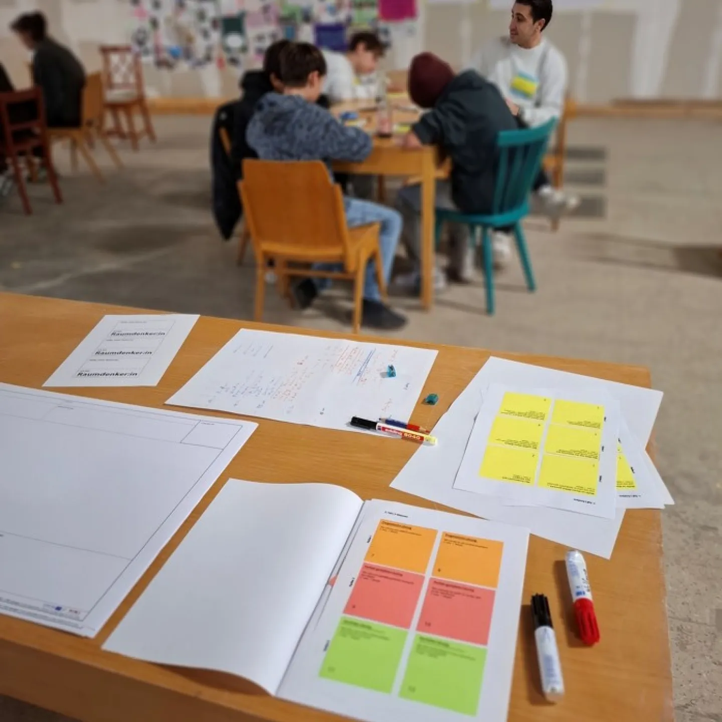 Blick auf einen Schreibtisch mit bunten Notizzetteln und Menschen, die an einem Tisch sitzen im Hintergrund