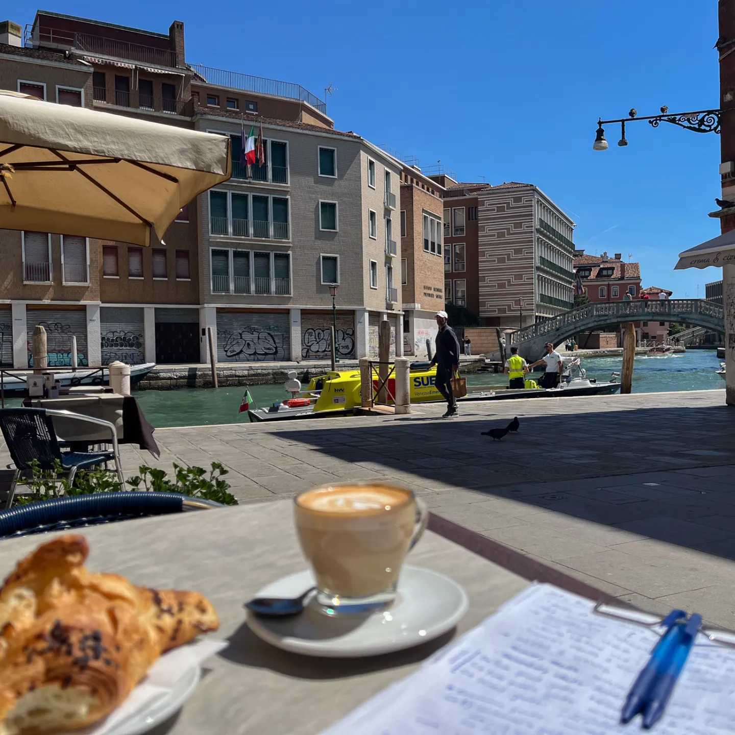 Coffee, croissant and notes on a table by a canal in Venice.