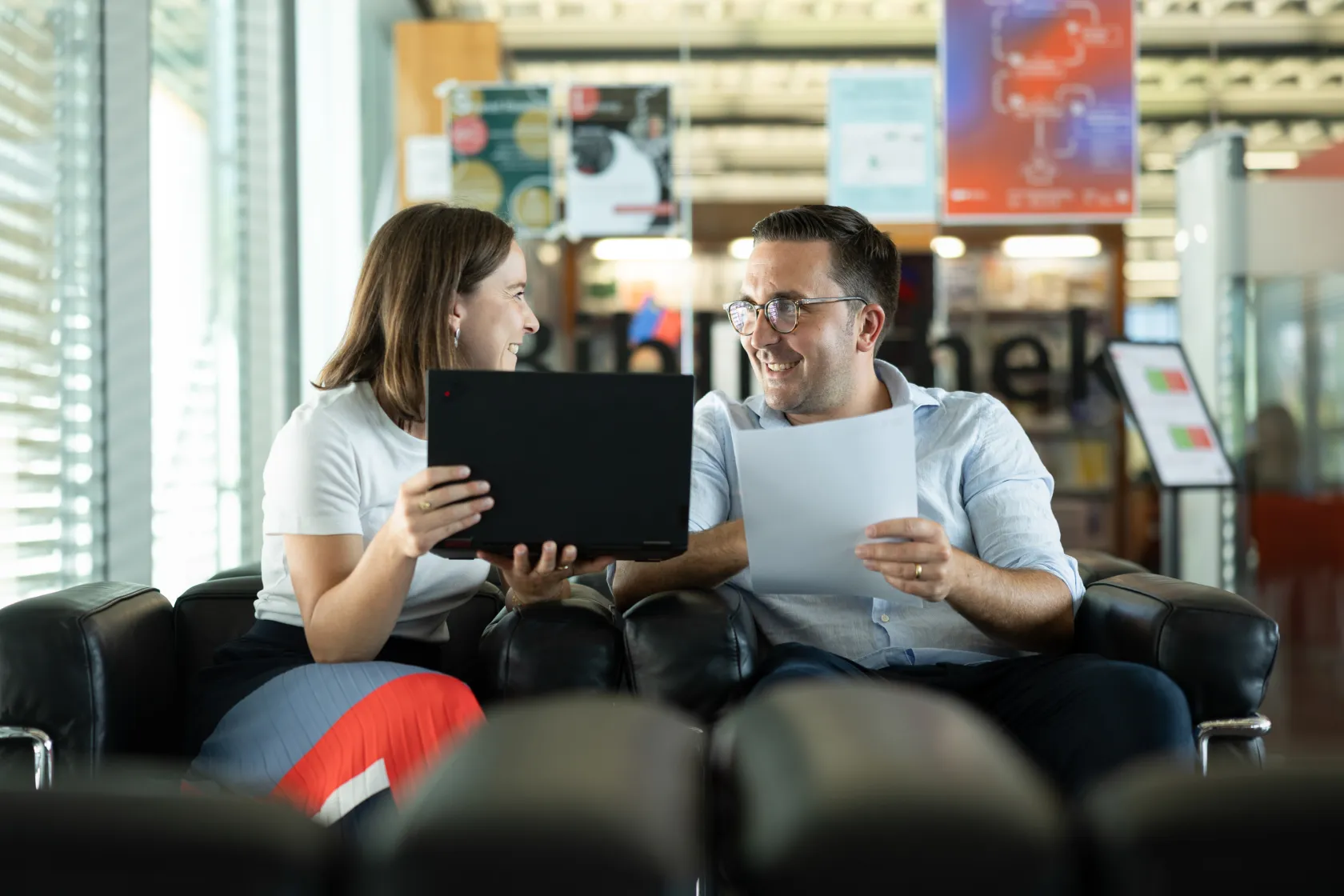 Two People Laughing During a Work Discussion with a Laptop and Documents in the Library Reading Area
