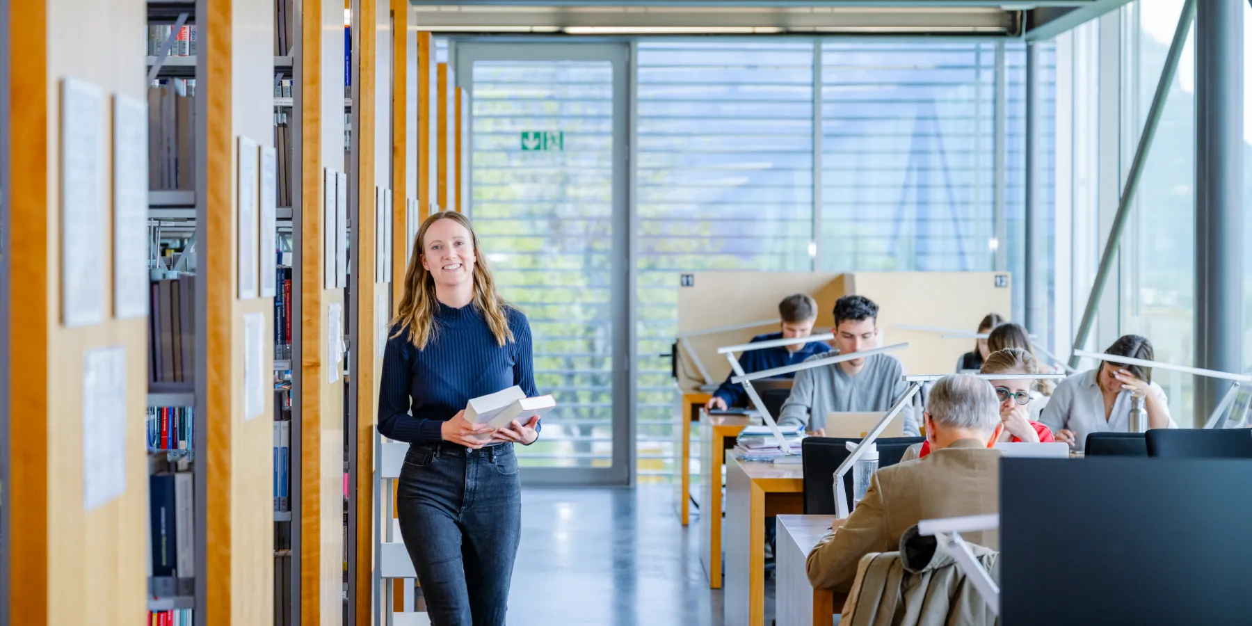 Studentin geht lächelnd mit Büchern durch die Regale der hellen Universitätsbibliothek der Universität Liechtenstein, im Hintergrund konzentriert arbeitende Personen an Einzelarbeitsplätzen.