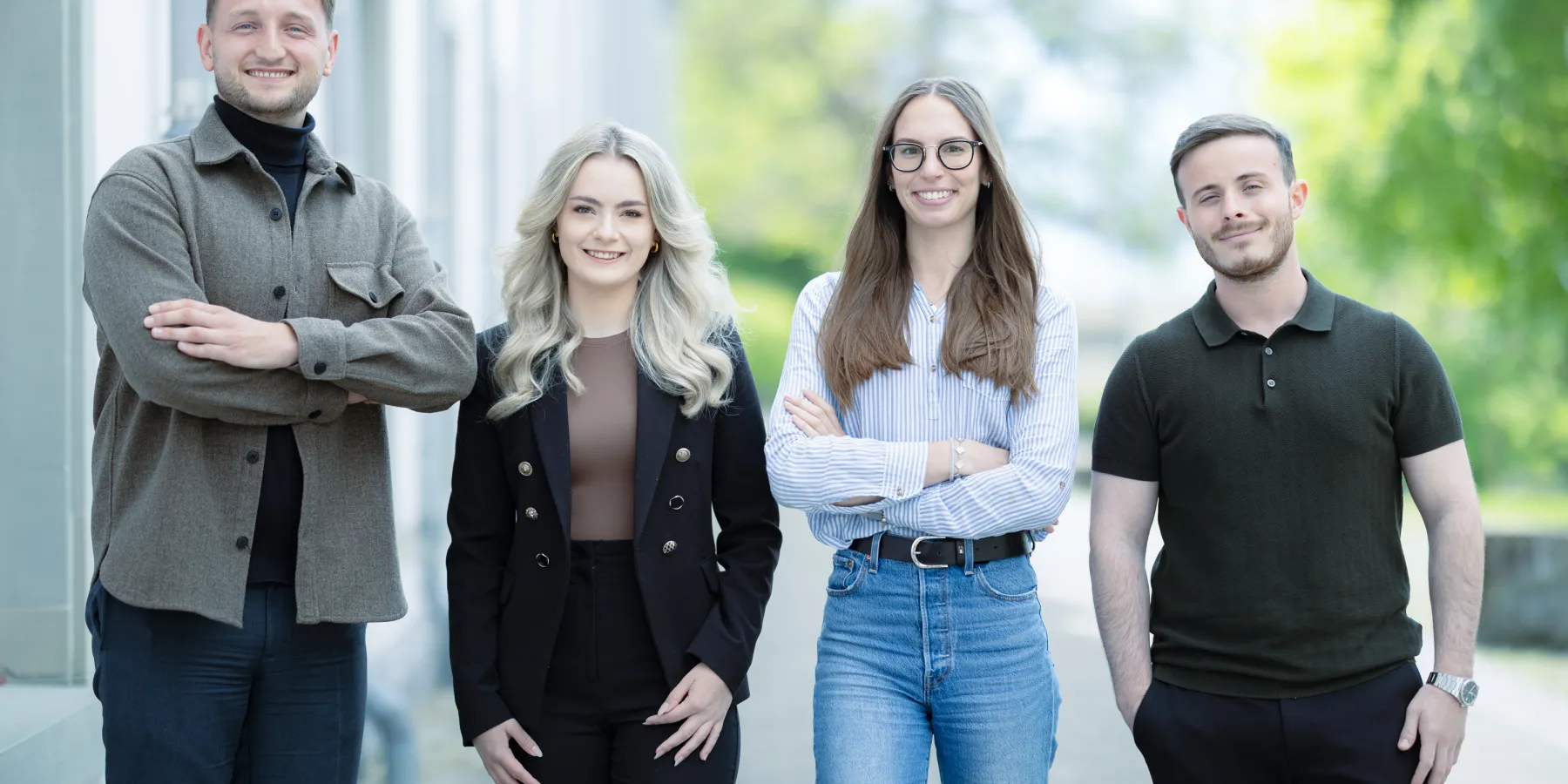 Vier Studierende im Masterstudiengang Information Systems der Universität Liechtenstein stehen nebeneinander auf einem Gehweg vor einem Universitätsgebäude und lächeln in die Kamera. Die Kleidung reicht von Business- bis Casual-Stil.