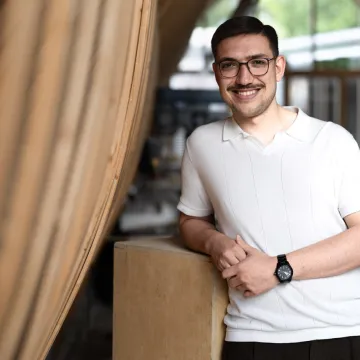 Junger Mann mit Brille und dunklem Haar, der in einer Werkstatt direkt in die Kamera blickt. 