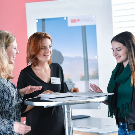 Drei Frauen im Gespräch an einem Stehtisch beim Infotag der Universität Liechtenstein, mit Informationsmaterialien und freundlicher Atmosphäre.